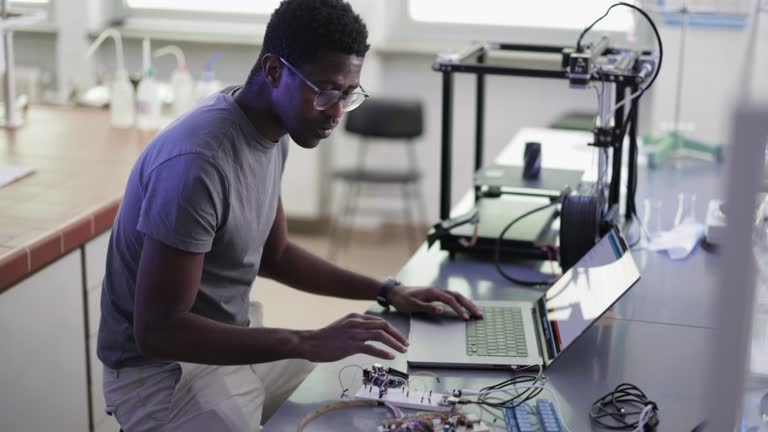 Young African-American technician sitting in the chair, thinking and repairing his 3D printer in the laboratory.